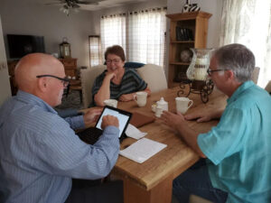 Gary Henicke sitting at a dining table reviewing a home sales pricing strategy on a tablet with smiling home sellers.